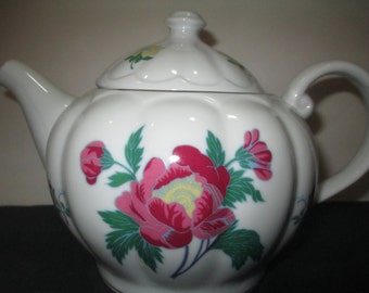 May include: White porcelain teapot with a floral design featuring pink, yellow, and green flowers. The teapot has a round body and a lid with a scalloped edge.