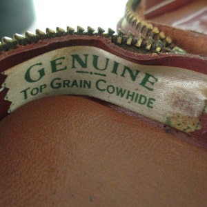 May include: Close-up of a brown leather zippered pouch with a white label that reads "GENUINE TOP GRAIN COWHIDE" in green lettering.