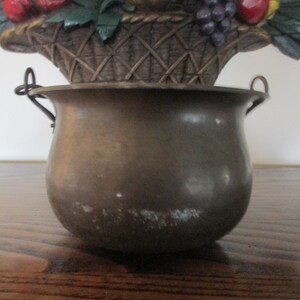 May include: A bronze-colored decorative pot with two small handles. The pot has a rounded shape and sits beneath a basket filled with faux fruit, including red apples and purple grapes. The basket is made of woven material.