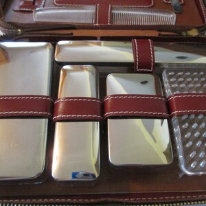 May include: A brown leather travel case with a zipper closure. The case is open and contains a set of six silver-toned grooming tools, including a comb, a razor, a brush, and three small containers.