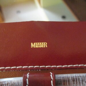May include: Close-up of a red leather bag with white stitching. The bag has a gold embossed label that reads "Meeker Made".