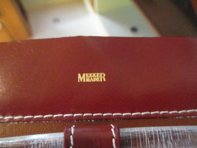 May include: Close-up of a red leather bag with white stitching. The bag has a gold embossed label that reads "Meeker Made".