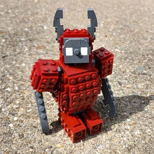 Peut inclure: Une figurine Lego rouge avec des accents gris, ressemblant à un robot avec des cornes. La figurine a un design en blocs et est assise sur une surface grise.
