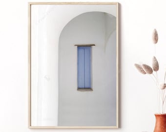 Può includere: Una fotografia incorniciata di un edificio bianco con una porta blu e un ingresso ad arco. La fotografia è appesa a una parete bianca sopra un tavolo con un vaso di fiori secchi e due vasi decorativi.