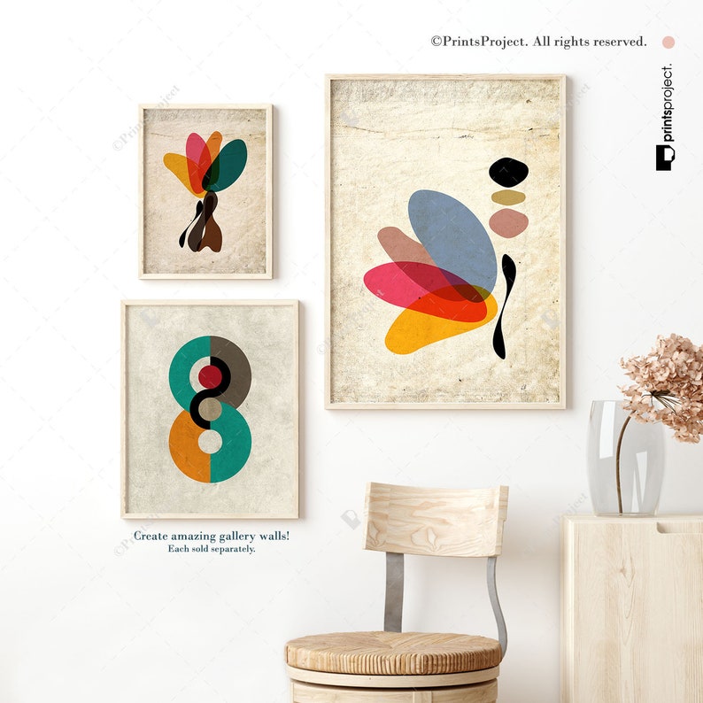 May include: Three abstract art prints with geometric shapes in shades of red, orange, yellow, blue, green, and brown. The prints are framed in a light wood frame and are displayed on a white wall.