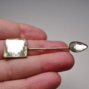 May include: A gold-toned stick pin with two clear faceted glass stones. The larger stone is square and the smaller stone is teardrop shaped.