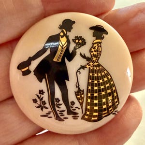 May include: A round, off-white brooch featuring black silhouettes of a man offering flowers to a woman. The man wears a suit with gold accents, and the woman holds an umbrella. The woman is wearing a checkered dress with gold accents. Decorative floral elements are at the bottom.