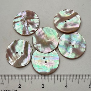 May include: Six iridescent, round buttons with two holes each. The buttons have a pearlescent sheen with colors of pink, green, and brown. A ruler is visible in the image, with measurements in inches.