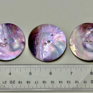 May include: Three iridescent purple and white shell buttons with two holes each. The buttons are approximately 2.5 cm in diameter and are arranged in a row on a white surface.