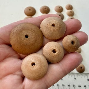 May include: A collection of unfinished wooden beads in various sizes, held in a hand. The beads are circular with a hole in the center, and range in color from light to medium brown. Some beads are in the background.