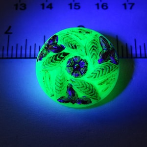May include: A green glass button with a floral design and a butterfly motif. The button is iridescent and glows under a black light.
