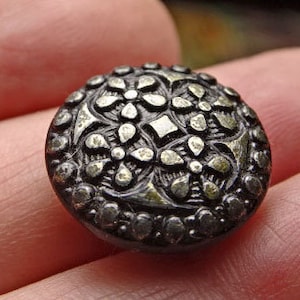 May include: A silver-toned button with a raised, intricate floral design. The button has a circular shape and a smooth, shiny surface.