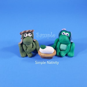 May include: A green frog wearing a brown and white hat and a green frog wearing a blue hat with a pink flower, both sitting beside a white egg in a brown basket. The frogs are a nativity scene.