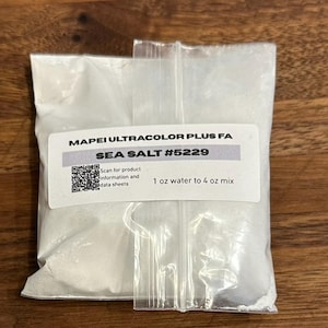May include: A light brown wooden craft stick, a clear plastic bag containing white Mapei Ultracolor Plus FA Sea Salt #5229 powder with a label stating 1 oz water to 4 oz mix, and a small, round, light brown paper cup with a white interior rim.