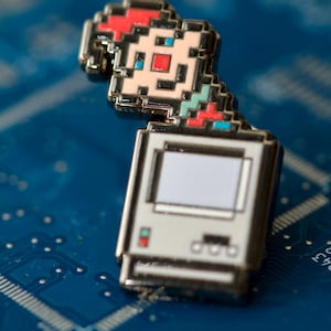 May include: A metal enamel pin featuring a pixelated computer monitor with a white screen and a small, colorful pixelated character figure perched on top. The character has red, light blue, and beige pixel details, while the computer is white.