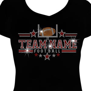 May include: A black V-neck t-shirt featuring a rhinestone graphic of a football, a goalpost, and stars. The design includes the text "TEAM NAME" and "FOOTBALL" in red and white crystals, creating a sparkling effect on the front of the shirt.
