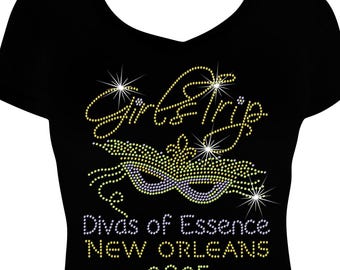 May include: A black V-neck t-shirt featuring a rhinestone design of a masquerade mask and the text Girl Trip, Divas of Essence, NEW ORLEANS, and 2025. The mask is decorated with yellow and purple rhinestones, while the text uses yellow and white.