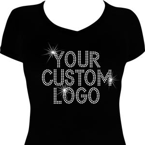 May include: Black V-neck t-shirt with the text "YOUR CUSTOM LOGO" in white rhinestones. The text is arranged in a stacked format with starburst accents. The shirt is a solid black color.
