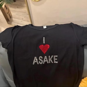 May include: A black short-sleeved t-shirt featuring a graphic design made of small rhinestones. The design consists of the letter I, a red heart shape, and the word ASAKE written in white rhinestones below the heart on the chest area.