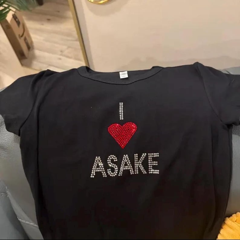 May include: A black short-sleeved t-shirt featuring a graphic design made of small rhinestones. The design consists of the letter I, a red heart shape, and the word ASAKE written in white rhinestones below the heart on the chest area.