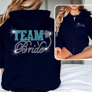 May include: Navy blue zip-up hoodie with the text "TEAM Bride" in sparkling rhinestones. The hoodie features a hood and a full-length zipper. A smaller image shows the name "Julia" in rhinestones.