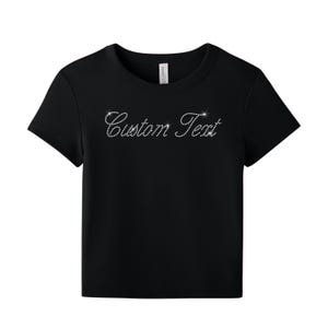 May include: A black short-sleeved t-shirt featuring the words Custom Text written in a cursive script font. The lettering is composed of small, reflective silver-toned rhinestones that create a sparkling effect against the dark fabric background.