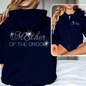 May include: Navy blue zip-up hoodie with the text "Mother of the Groom" in sparkling rhinestones. The design includes a ring graphic. A smaller image shows a similar hoodie with the name "Julia".