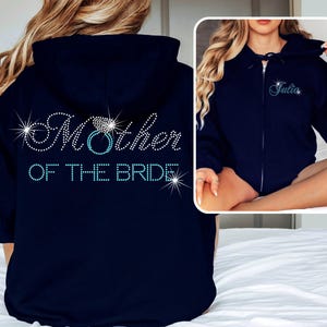 May include: Navy blue zip-up hoodie with the text "Mother of the Bride" in sparkling rhinestones on the back. The design includes a ring graphic. A smaller image shows a similar hoodie with the name "Julia".