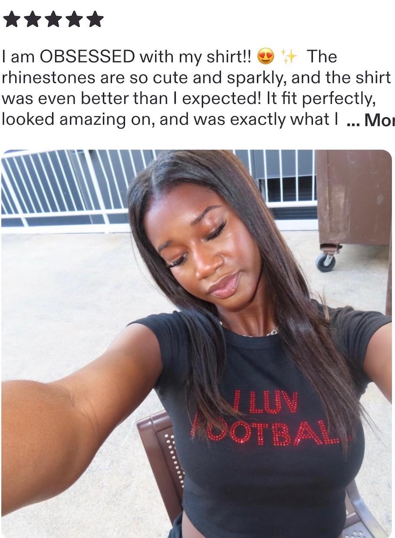 May include: A black short-sleeved t-shirt featuring the red rhinestone text I LUV FOOTBALL on the chest. The shirt has a crew neckline and a fitted silhouette. The red rhinestones are arranged to form the letters of the text clearly.