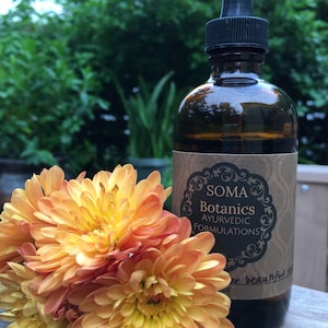 May include: A brown glass bottle with a dropper top and a label that reads "Soma Botanics Ayurvedic Formulations". The bottle is sitting on a wooden surface next to a bouquet of orange and yellow flowers.