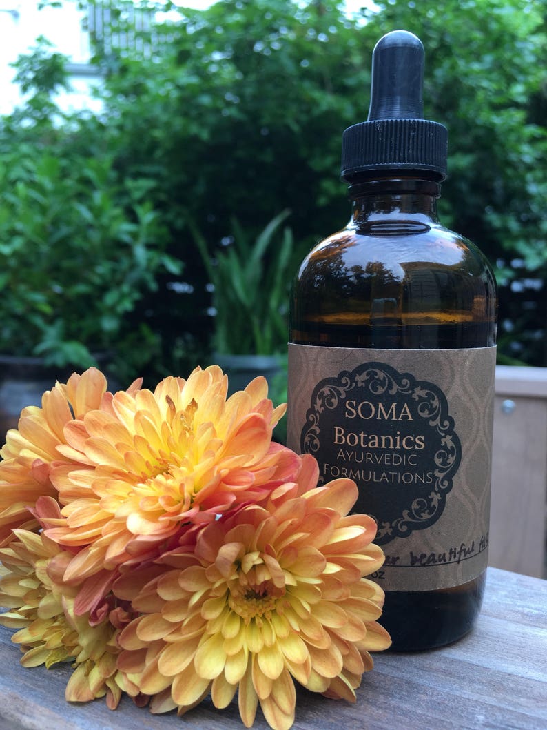 May include: A brown glass bottle with a dropper top and a label that reads "Soma Botanics Ayurvedic Formulations". The bottle is sitting on a wooden surface next to a bouquet of orange and yellow flowers.