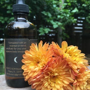 May include: A dark amber glass bottle with a black dropper top and a label that reads "Formulated with an essential oil blend, specifically for you: your signature body bouquet" and "&copy;2015 SOMAbota Miami, FL 33141 Somabotanics@gmail.com". The bottle is sitting on a wooden surface with a bouquet of orange flowers.