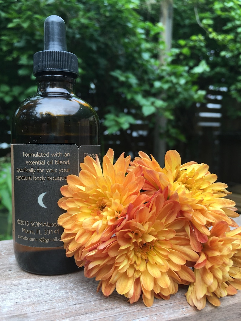 May include: A dark amber glass bottle with a black dropper top and a label that reads "Formulated with an essential oil blend, specifically for you: your signature body bouquet" and "&copy;2015 SOMAbota Miami, FL 33141 Somabotanics@gmail.com". The bottle is sitting on a wooden surface with a bouquet of orange flowers.