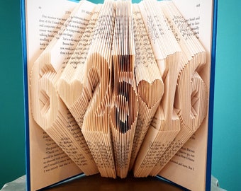 May include: A book art sculpture with the words "LOVE" folded into the pages. The book is bound in blue and has white pages. The sculpture is on a wooden surface.