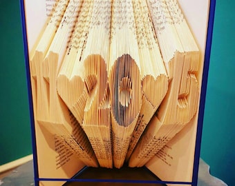 May include: A book sculpture with the numbers "11 20 15" cut into the pages. The book is open, with the pages fanned out to display the numbers and heart shapes. The book has a blue cover and is held together with a blue elastic band.
