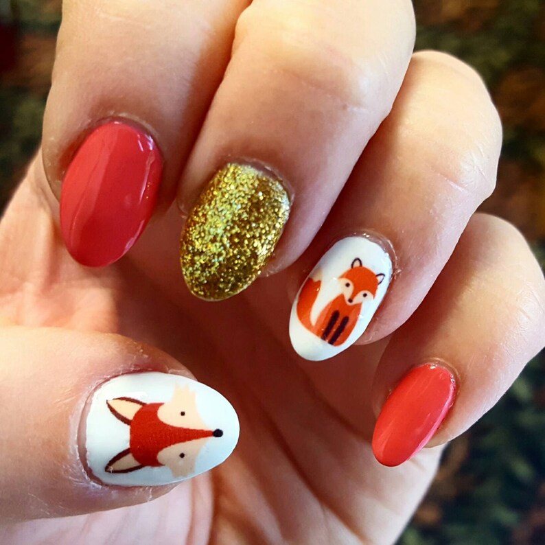 May include: Close-up of a hand with five fingernails painted with different designs. The thumb and ring finger are painted a bright red. The index finger is painted with gold glitter. The middle finger and pinky finger are painted white with a small orange fox design on each.