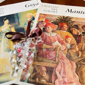 May include: Two art books with brown ribbon tied around them. The book on the left is titled "I Maestri del Colore Goya" and the book on the right is titled "I Maestri del Colore Mantegna".