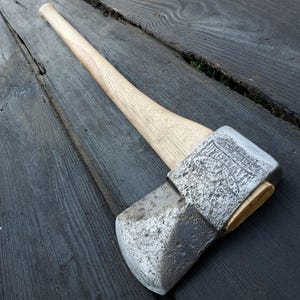 May include: A vintage axe with a wooden handle and a steel head. The axe head has a textured surface and is engraved with decorative elements. The wooden handle is light-colored and tapers towards the end. The axe is designed for chopping wood.
