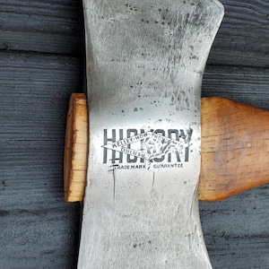 May include: Close-up of a vintage double-bit axe with a wooden handle. The axe head is silver with the text "HICKORY" and "KELLEY-HOW-THOMSON DULUTH MINN" printed on it. The axe is set against a dark wood background.