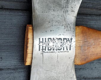 Puede incluir: Primer plano de un hacha de doble filo vintage con mango de madera. La cabeza del hacha es plateada con el texto "HICKORY" y "KELLEY-HOW-THOMSON DULUTH MINN" impreso. El hacha está sobre un fondo de madera oscura.