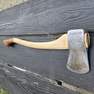 May include: A metal axe head with a curved wooden handle. The axe head features a circular stamp that reads HIGHEST QUALITY AMERICANAX GLASSPORT PA. The metal surface shows signs of wear and age, while the wooden handle has a smooth, light brown finish.