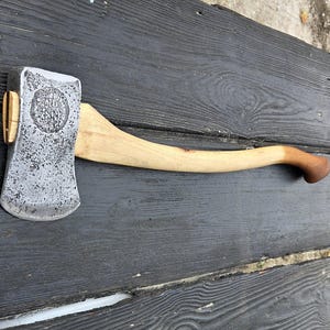 May include: A metal axe head with a textured, weathered finish and a circular stamp reading OLD HICKORY, attached to a curved, light-colored wooden handle that transitions to a darker brown tone at the grip end, featuring a small wooden wedge.