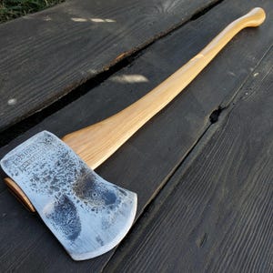 May include: Axe with a light-colored wooden handle and a silver-colored head. The axe head has a textured surface and is marked with text. The axe is lying on a dark wooden surface.
