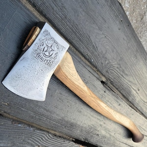 Könnte beinhalten: Ein Axtkopf aus Metall mit einem geschwungenen Holzstiel. Der Axtkopf zeigt ein eingestanztes kreisförmiges Logo mit einem Stern in der Mitte und dem Text THE DORIAN QUALITY AXE. Die Metalloberfläche hat ein strukturiertes, verwittertes Finish.