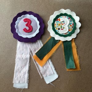 May include: Two handmade fabric award rosettes featuring the number 3. The left rosette has a purple felt base, a light purple center, and a red felt number 3 with white lace ribbons. The right rosette has a white felt base, a floral center, and a white felt number 3 with green and mustard yellow ribbons.