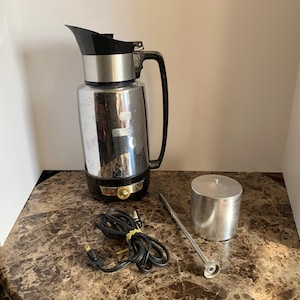 May include: A silver and black electric coffee pot with a cord and a separate silver filter. The coffee pot has a black handle and a gold accent.