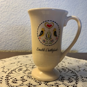 May include: White ceramic mug with a black, red, yellow, and green design of two birds and a heart. The mug has a footed base and says "Double Dittlefink" on the front.