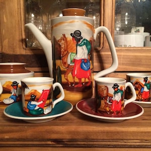 May include: A set of colorful ceramic teacups and a teapot with a brown lid. The cups and teapot feature hand-painted scenes of people and animals in a desert landscape.