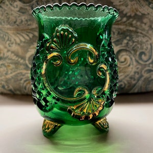 May include: A small, green glass vase with gold accents and a scalloped rim. The vase has three gold feet and a decorative design on the front.