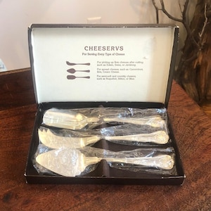 May include: A set of four silver cheese knives in a brown box with the text "CHEESERVS For Serving Every Type of Cheese" on the lid. The knives are wrapped in plastic and include a spreader, a knife for soft cheese, a knife for semi-hard cheese, and a knife for hard cheese.